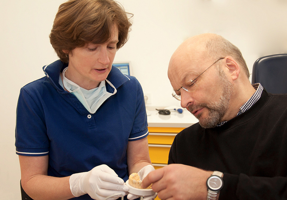 Lüdemannstraße 7A. Zahnärztin Dr. Reinhardt zeigt einem Patienten Zahnersatz aus Vollkeramik. 
